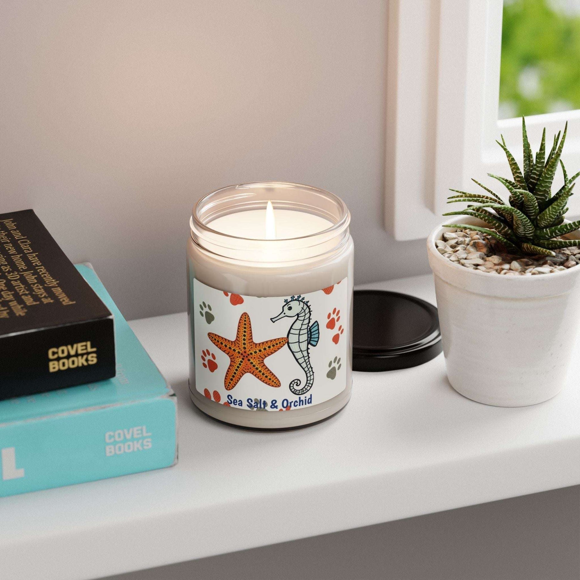 Scented soy candle in glass jar with sea salt and orchid aroma, on a shelf next to books and a plant.